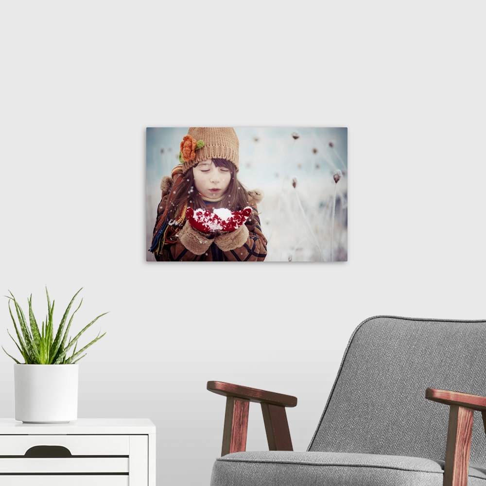 A modern room featuring A young girl in winter clothes blows on a handful of snow.