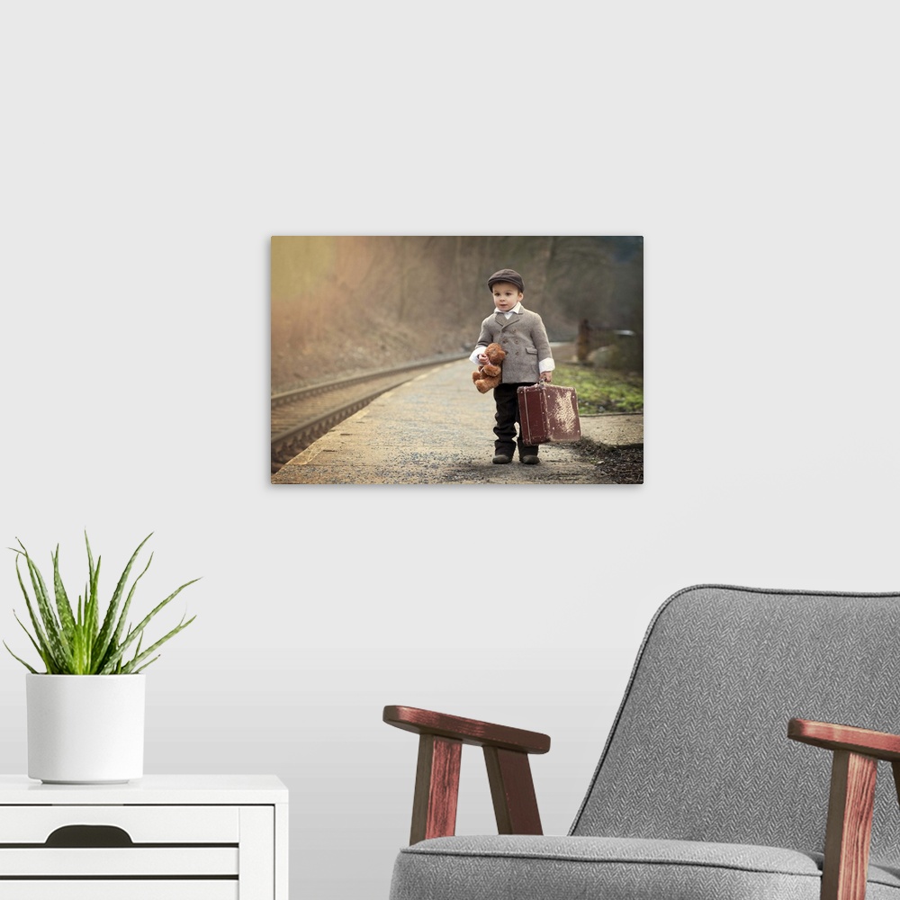 A modern room featuring A young boy holding a teddy bear and a suitcase waits near train tracks.