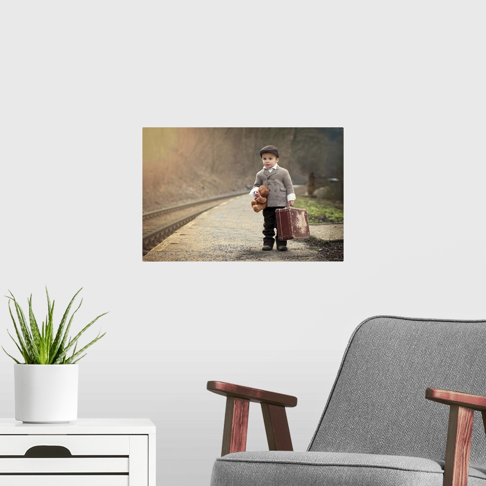 A modern room featuring A young boy holding a teddy bear and a suitcase waits near train tracks.