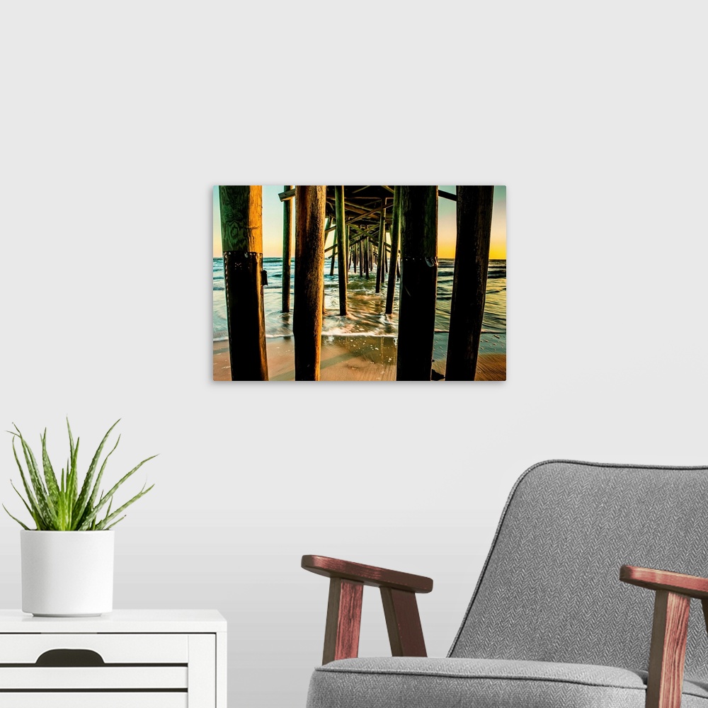 A modern room featuring A photograph looking through the underside of a long pier out over the ocean. The sky glowing wit...