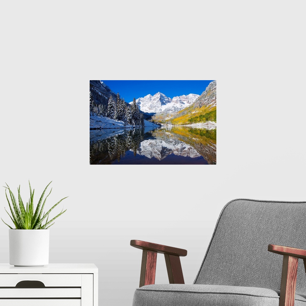 A modern room featuring A snow covered mountain range with tall trees in front reflects beautifully into a body of water.