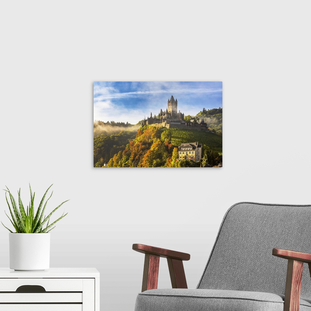 A modern room featuring Large medieval castle on a colorful tree hillside with fog, blue sky, and cloud, Cochem, Germany.