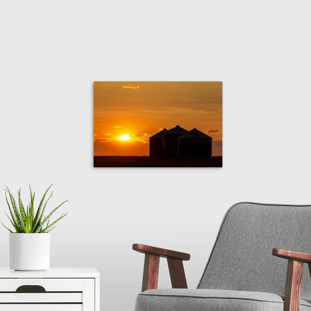 A modern room featuring Silhouette of large metal grain bins at sunrise with orange sun rising over clouds; Alberta, Canada