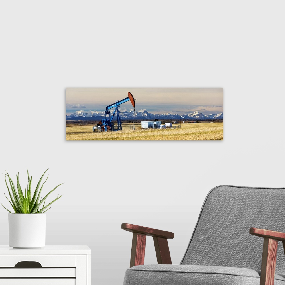 A modern room featuring Panorama of a colourful pump jack in cut canola stubble field with snow capped mountains, blue sk...