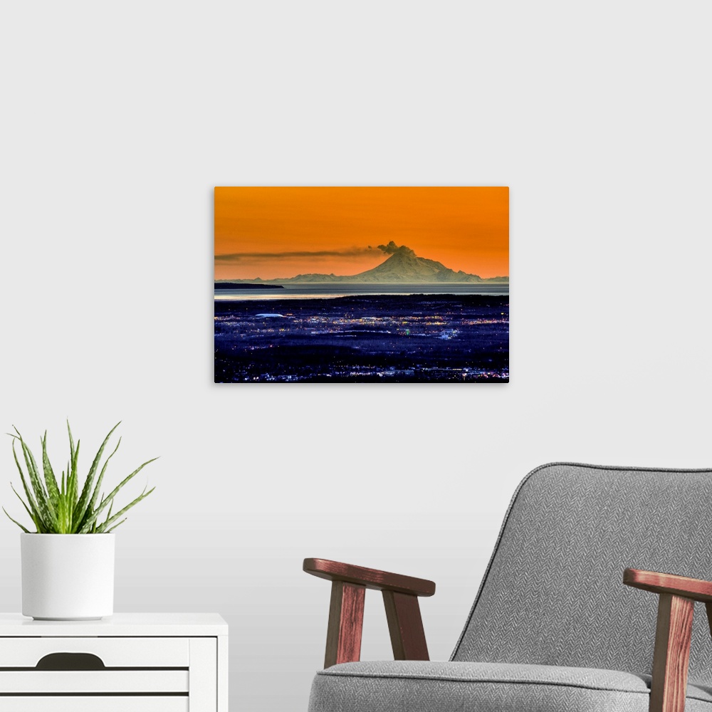 A modern room featuring Ash billows out of an active volcano which looms over a city in the evening in the Aleutian mount...