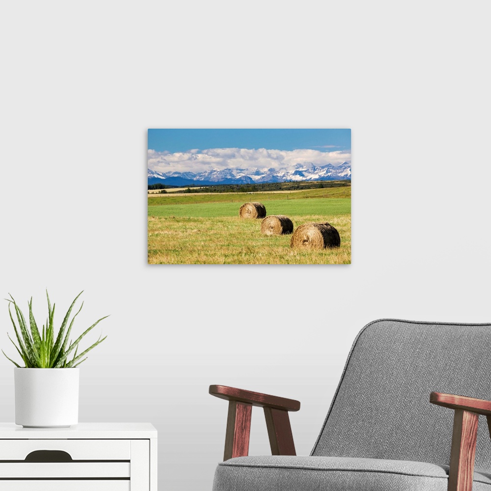 A modern room featuring Three hay bales in a field with mountains in the background slightly snow covered and cloudy with...