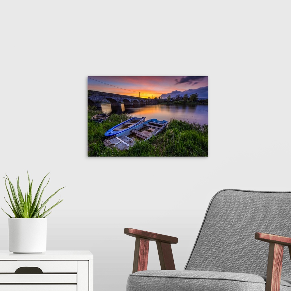 A modern room featuring Two small wooden boats on the bank of the Shannon river in front of a stone bridge at sunset in s...