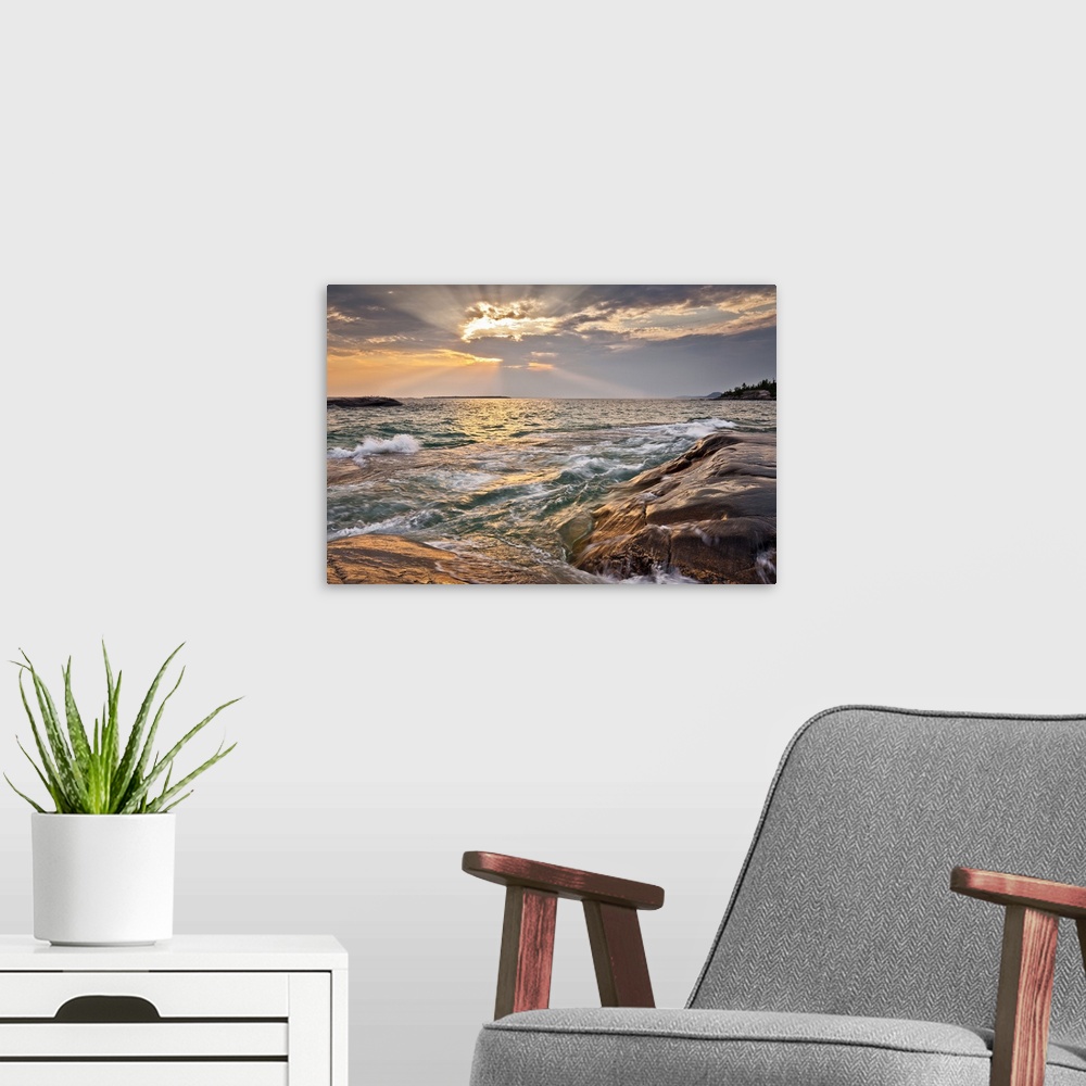 A modern room featuring Burst of sun rays peeking out behind a cloud as waves lap on to a rocky shore.