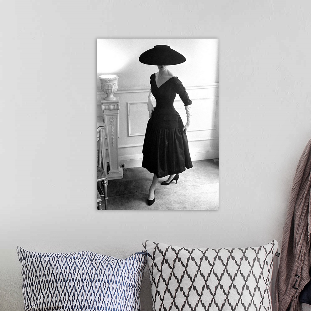 A bohemian room featuring A model wearing a black cocktail dress and a black velvet wide-brimmed hat, designed by Ronald Pa...