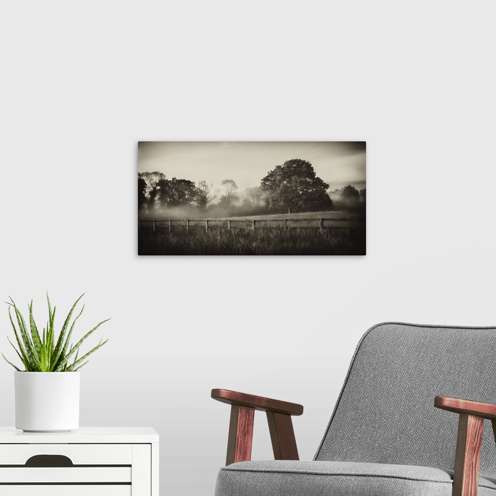 A modern room featuring A horizontal photograph of a country field with a wooden fence and mist over trees in the distance.