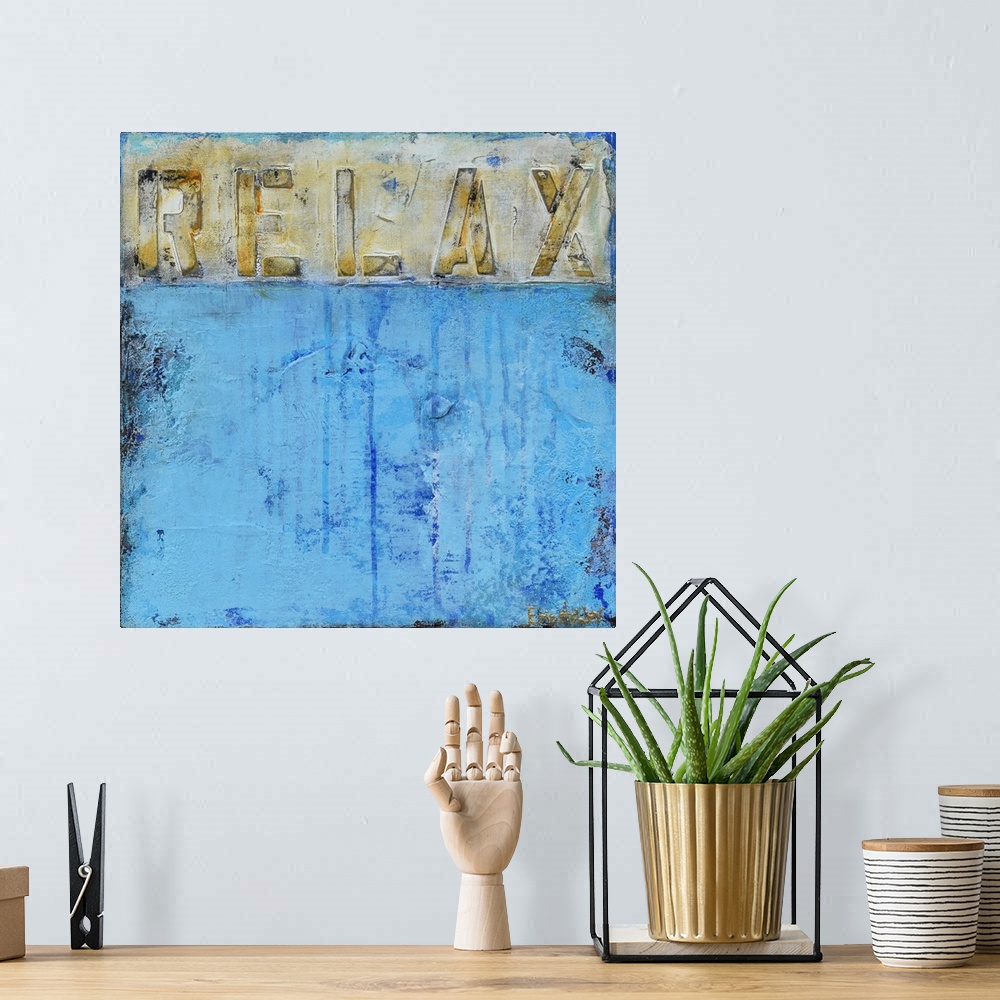 A bohemian room featuring "Relax" written in gray, gold, and white across the top of a square painting with a light blue ab...