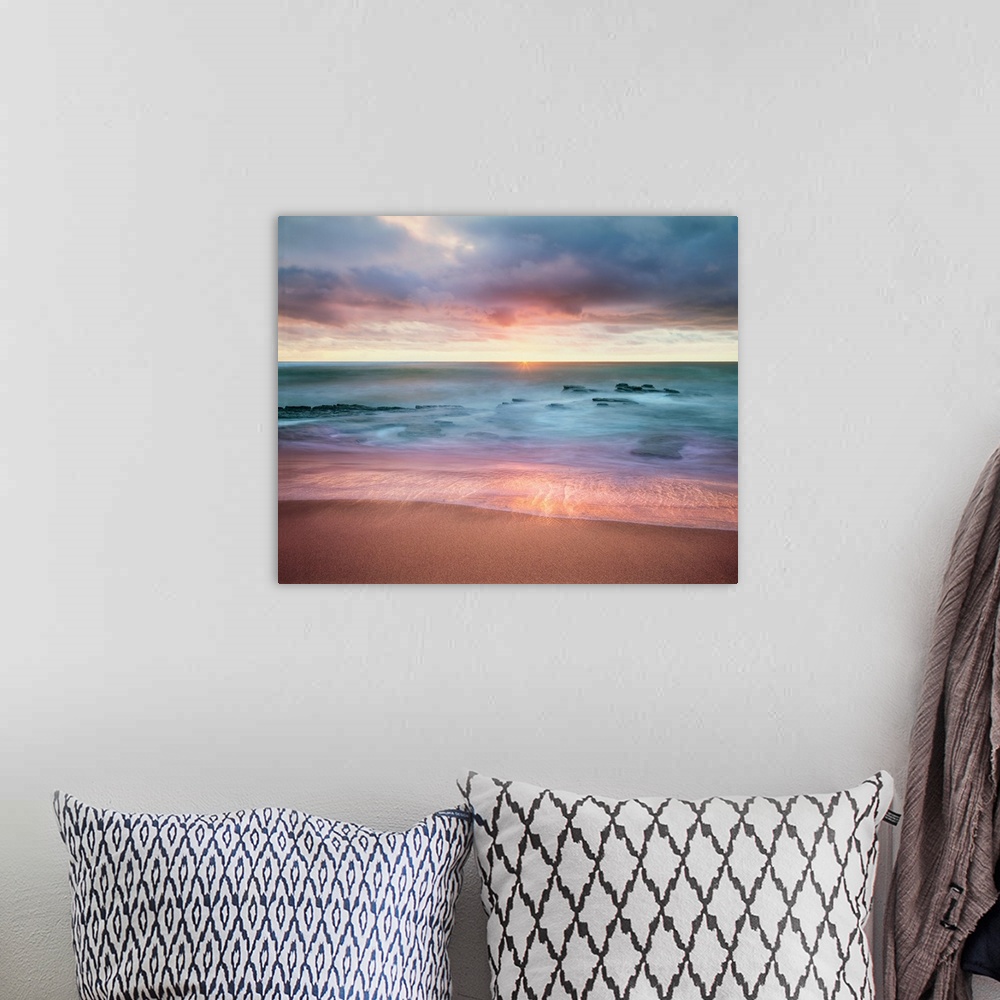 A bohemian room featuring Colorful long exposure photograph a rocky ocean shore with the sun on the horizon in shades of bl...
