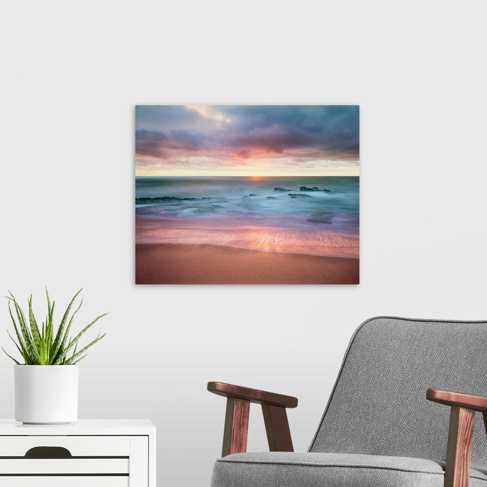 A modern room featuring Colorful long exposure photograph a rocky ocean shore with the sun on the horizon in shades of bl...