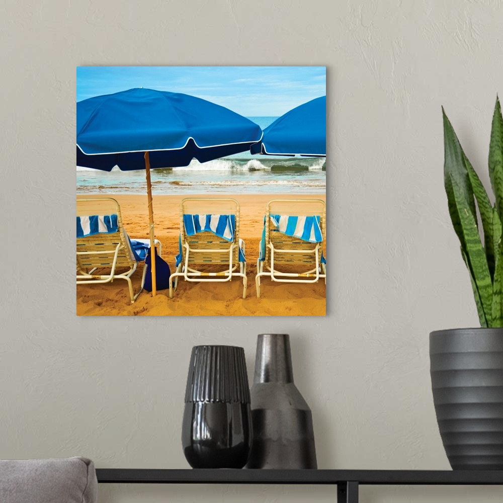 A modern room featuring Square photograph of three beach chairs with blue umbrellas lined up right in front of the ocean.