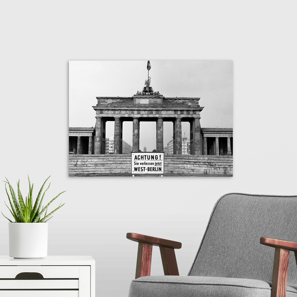 A modern room featuring The Brandenburg Gate in East Berlin. In front, a sign in German warns of the impending border bet...