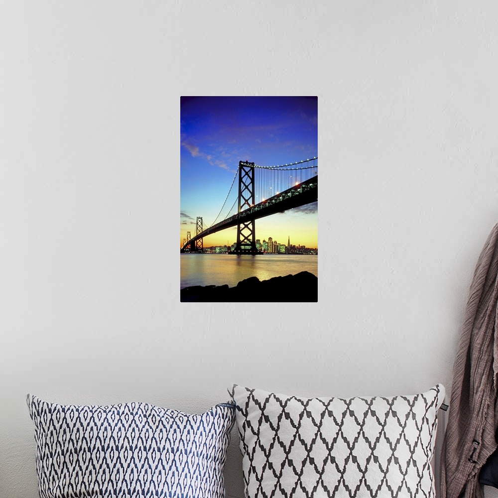 A bohemian room featuring The bay bridge is silhouetted under a sunset sky with the skyline in the distance lit up.