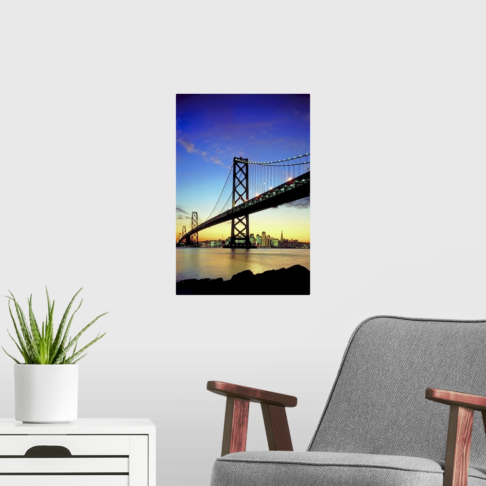 A modern room featuring The bay bridge is silhouetted under a sunset sky with the skyline in the distance lit up.