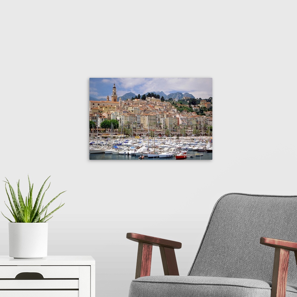 A modern room featuring French harbor full of boats with sails down, near a group of waterfront buildings nestled togethe...