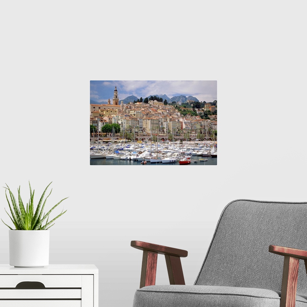 A modern room featuring French harbor full of boats with sails down, near a group of waterfront buildings nestled togethe...