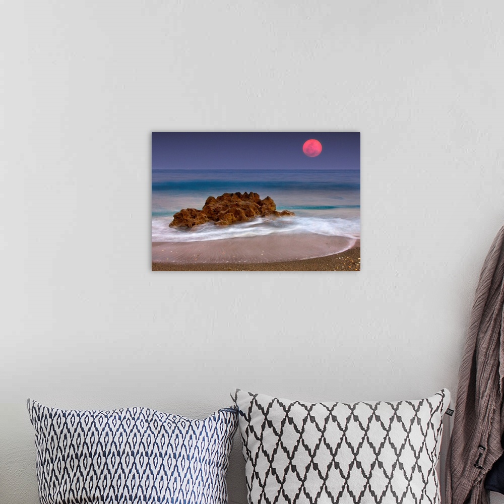 A bohemian room featuring A unique rock formation sits in the ocean water which is photographed under a deep red moon.
