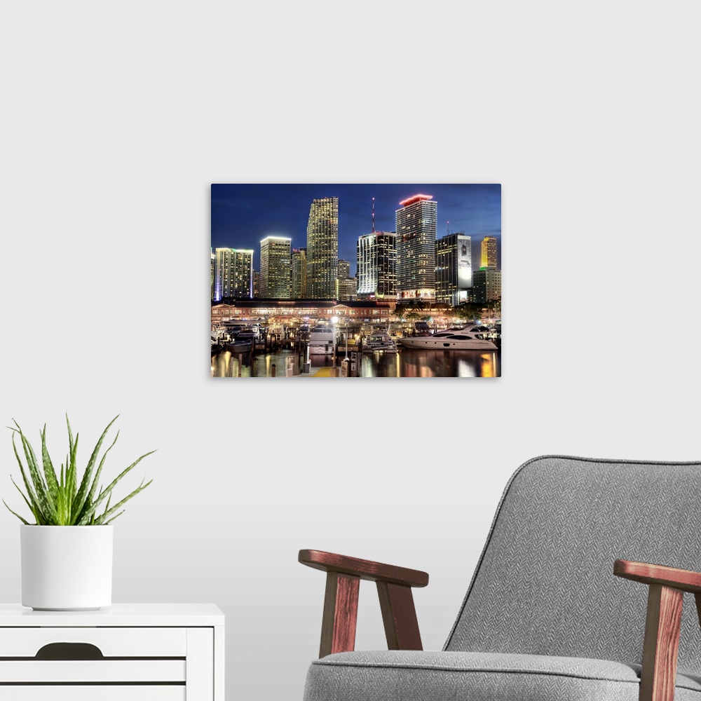 A modern room featuring Photograph of the nighttime lit up Miami skyline down at the harbor with boats parked in their wa...