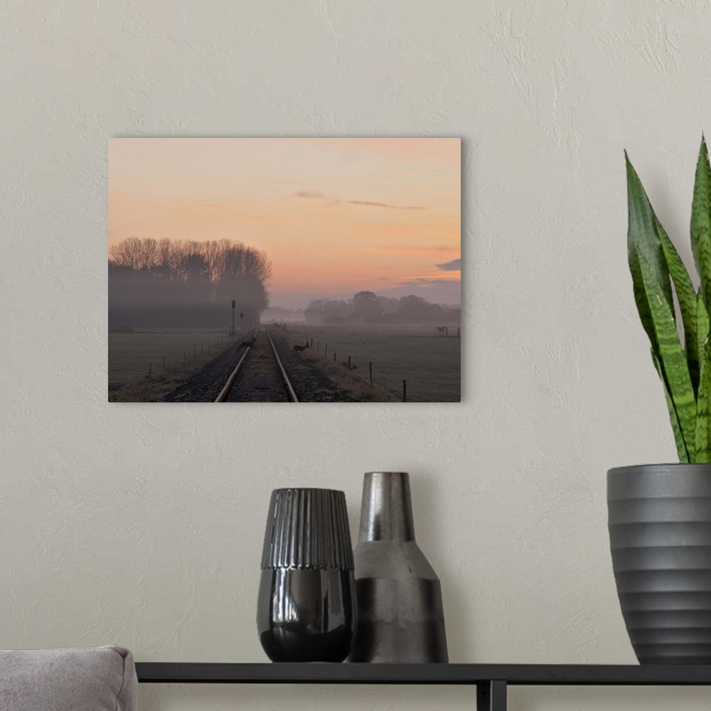 A modern room featuring Misty morning sunrise by single railway track with three deer crossing track and farm in distance.