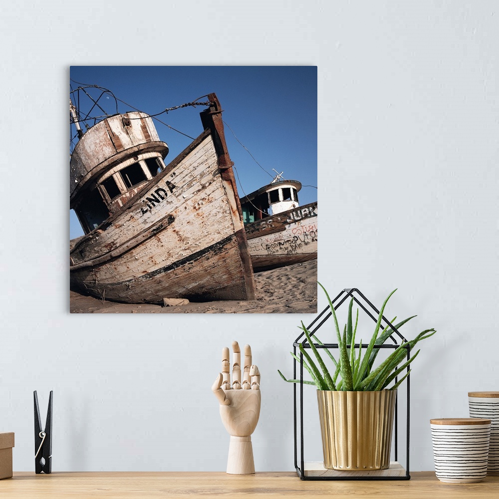 A bohemian room featuring old neglected boats sit beached on sand under a blue sky