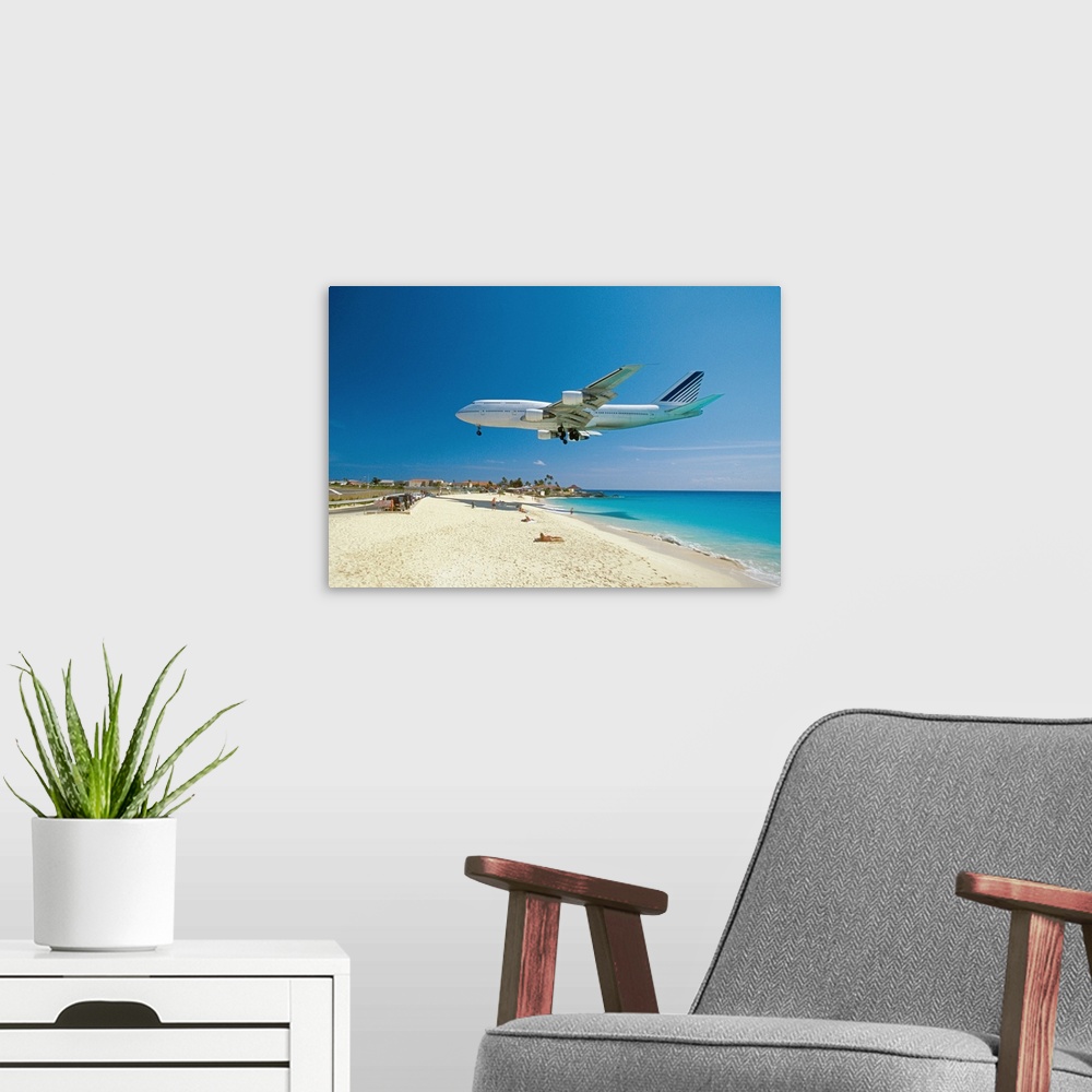 A modern room featuring This is a landscape photograph of a passenger jet flying low over a tropical beach preparing to l...
