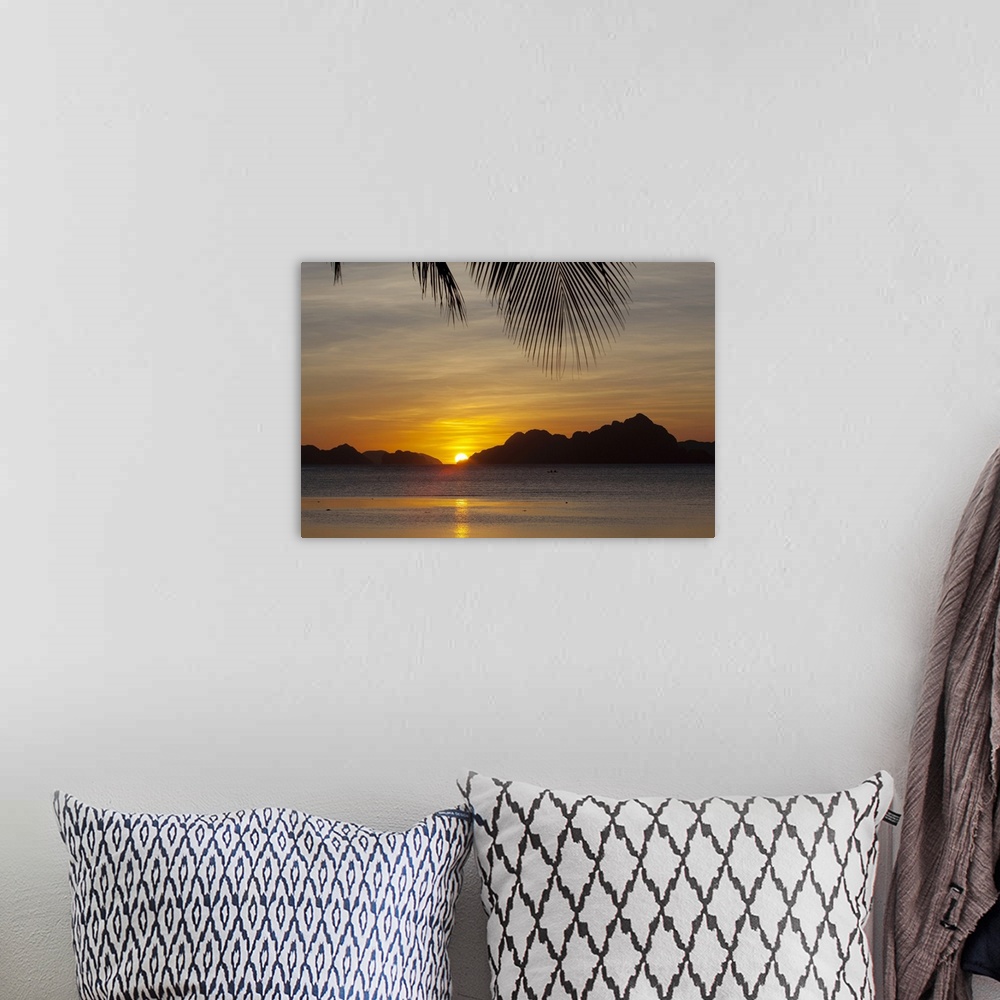 A bohemian room featuring Large, landscape photograph of the sun setting behind islands, a large palm branch hangs overhead...