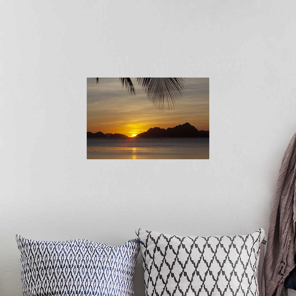 A bohemian room featuring Large, landscape photograph of the sun setting behind islands, a large palm branch hangs overhead...