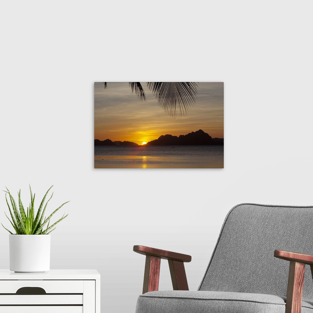 A modern room featuring Large, landscape photograph of the sun setting behind islands, a large palm branch hangs overhead...