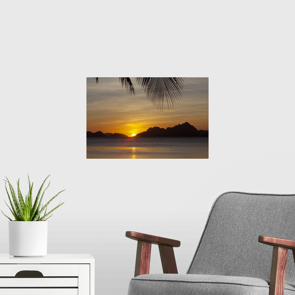 A modern room featuring Large, landscape photograph of the sun setting behind islands, a large palm branch hangs overhead...