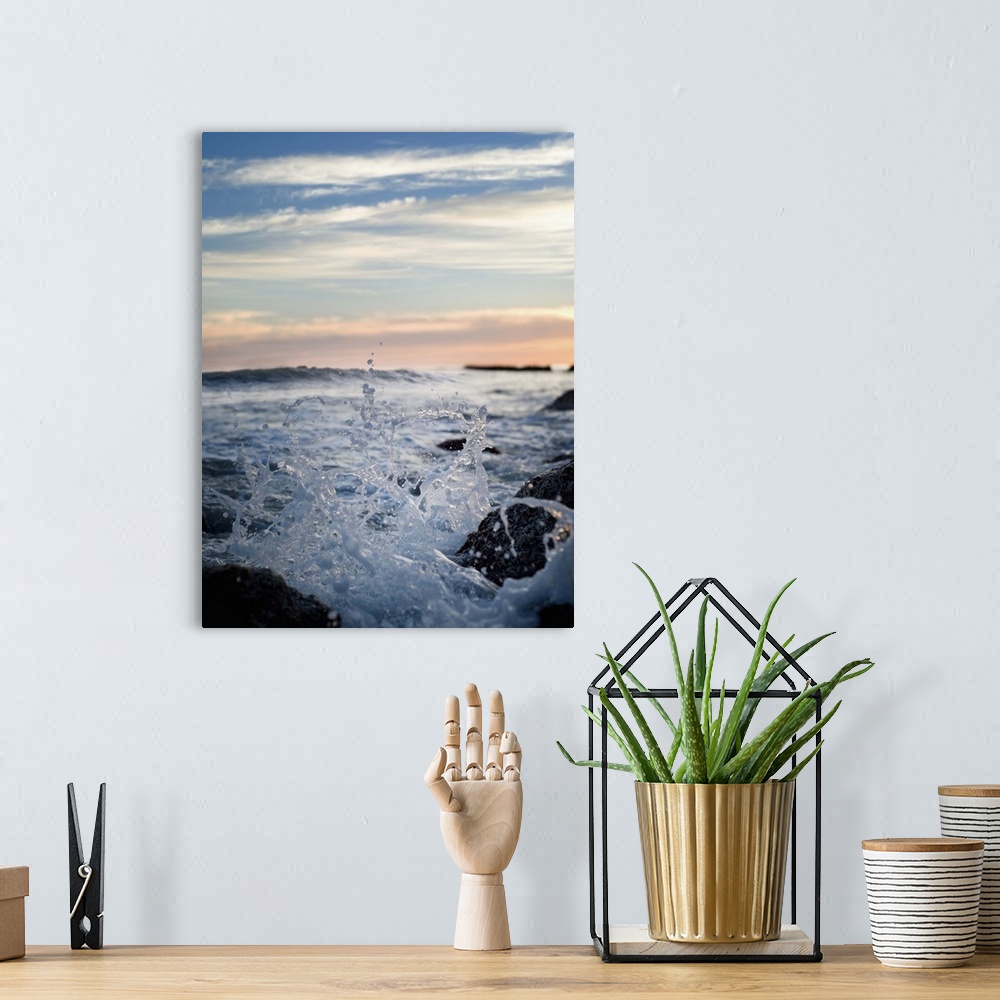 A bohemian room featuring Wave splashing against rocks on shore in the evening.  Sun is setting in background.
