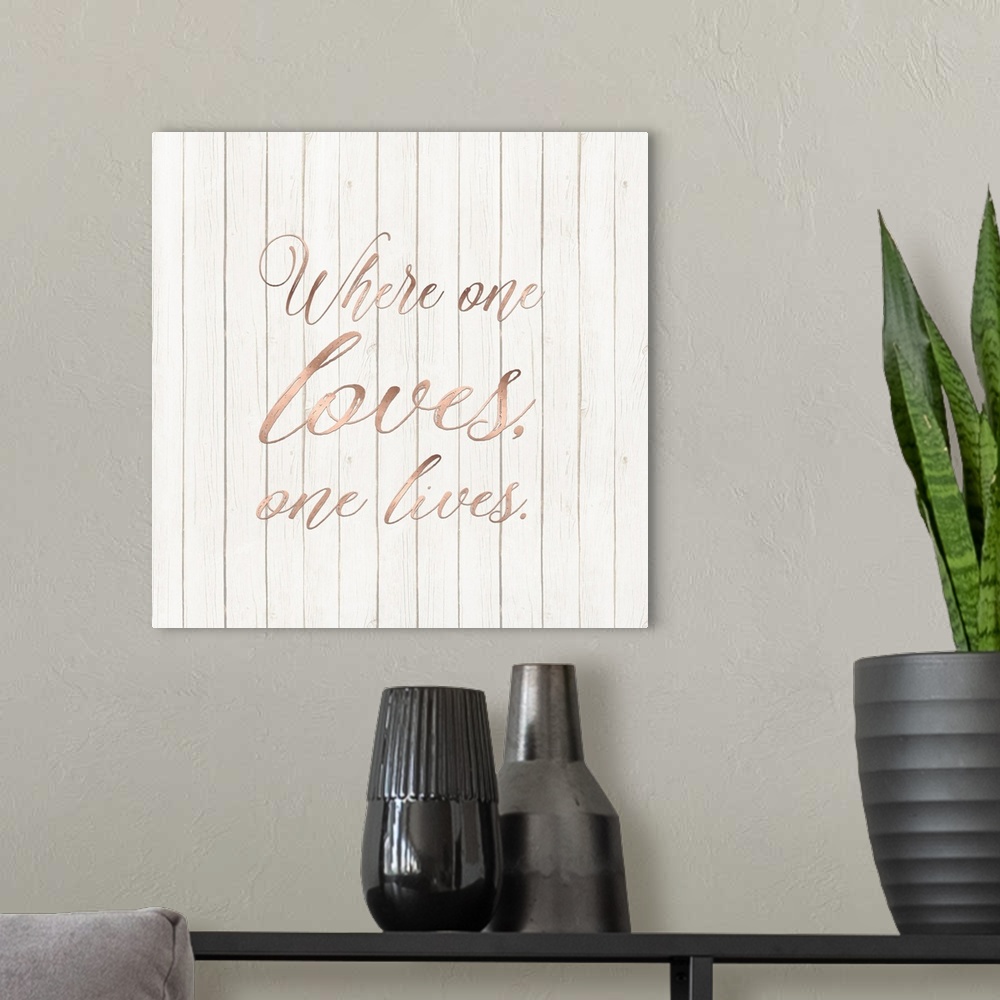 A modern room featuring "Where One Loves, One Lives." written in rose gold on a wood paneled background.