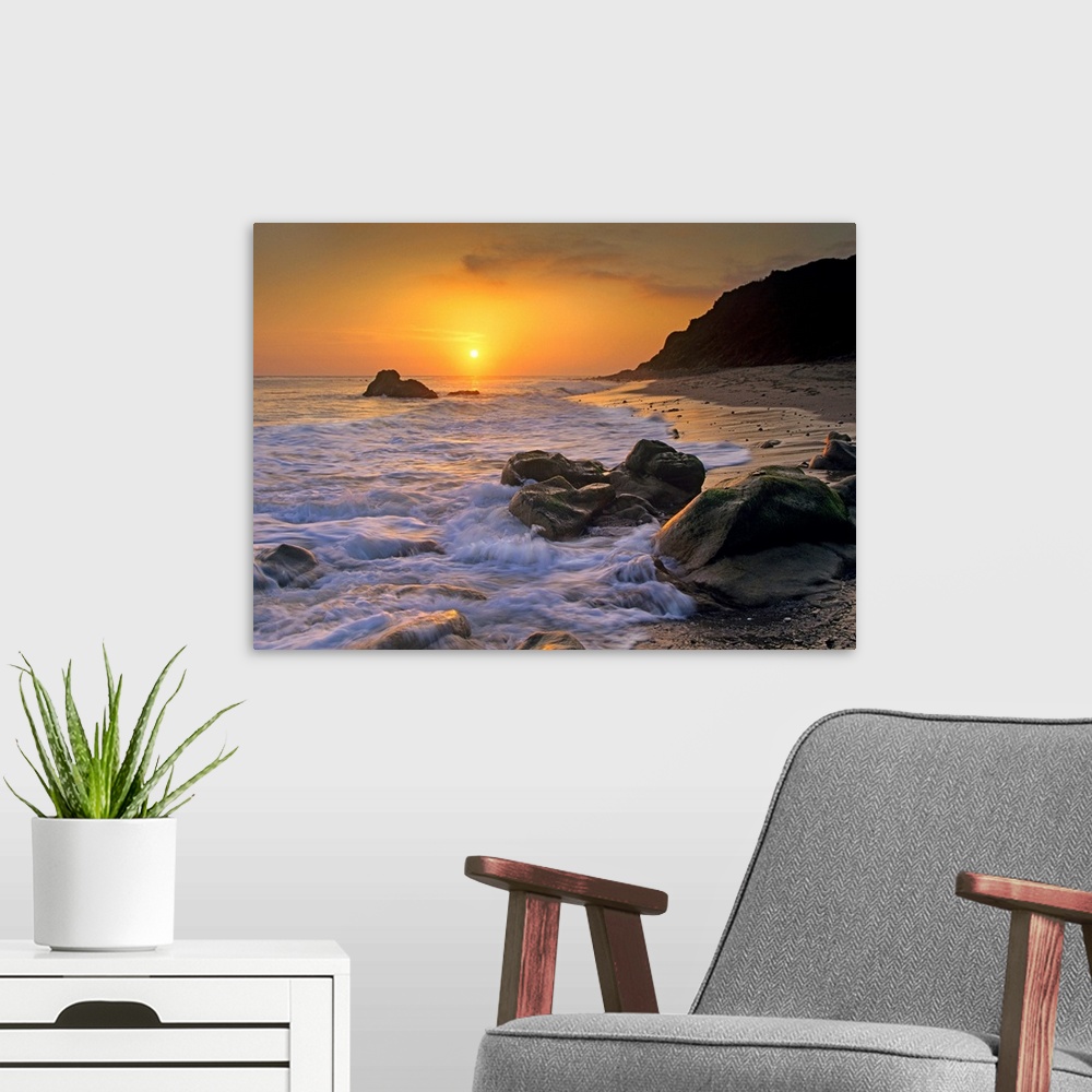 A modern room featuring Large landscape photograph of a golden sunset over the rocky shoreline at Leo Carrillo State Beac...