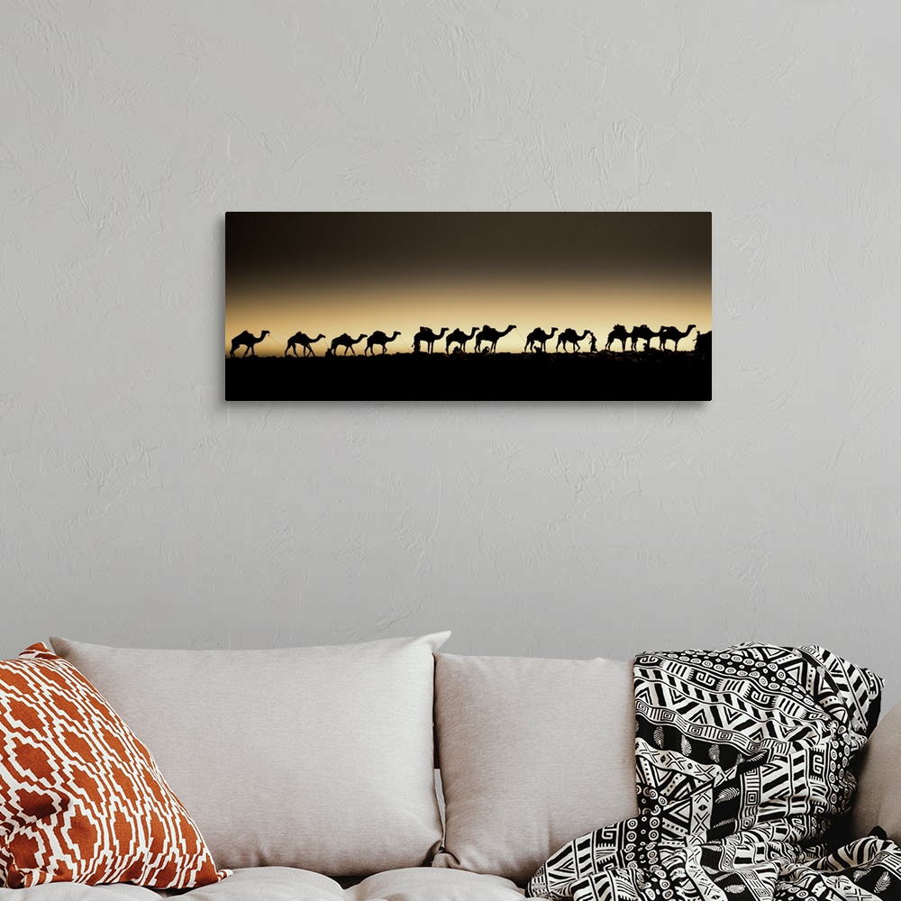 A bohemian room featuring A herd of camels in a single-file line, silhouetted against the setting sun.