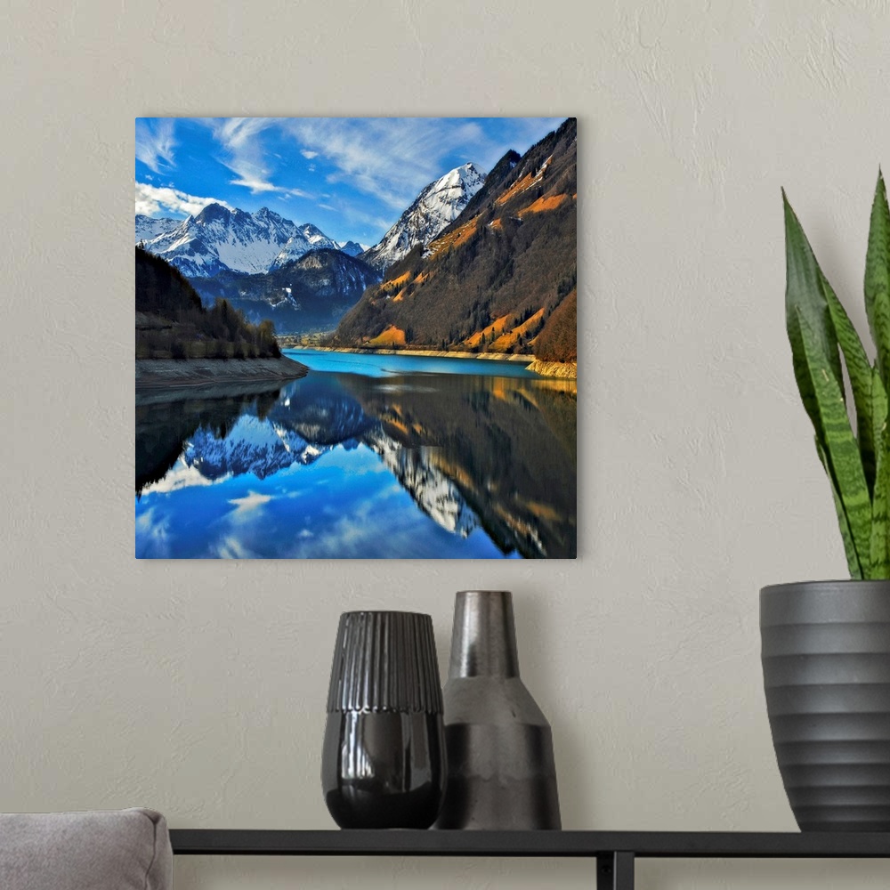 A modern room featuring Square photograph on a big wall hanging of snow covered mountains beneath a blue sky, reflecting ...