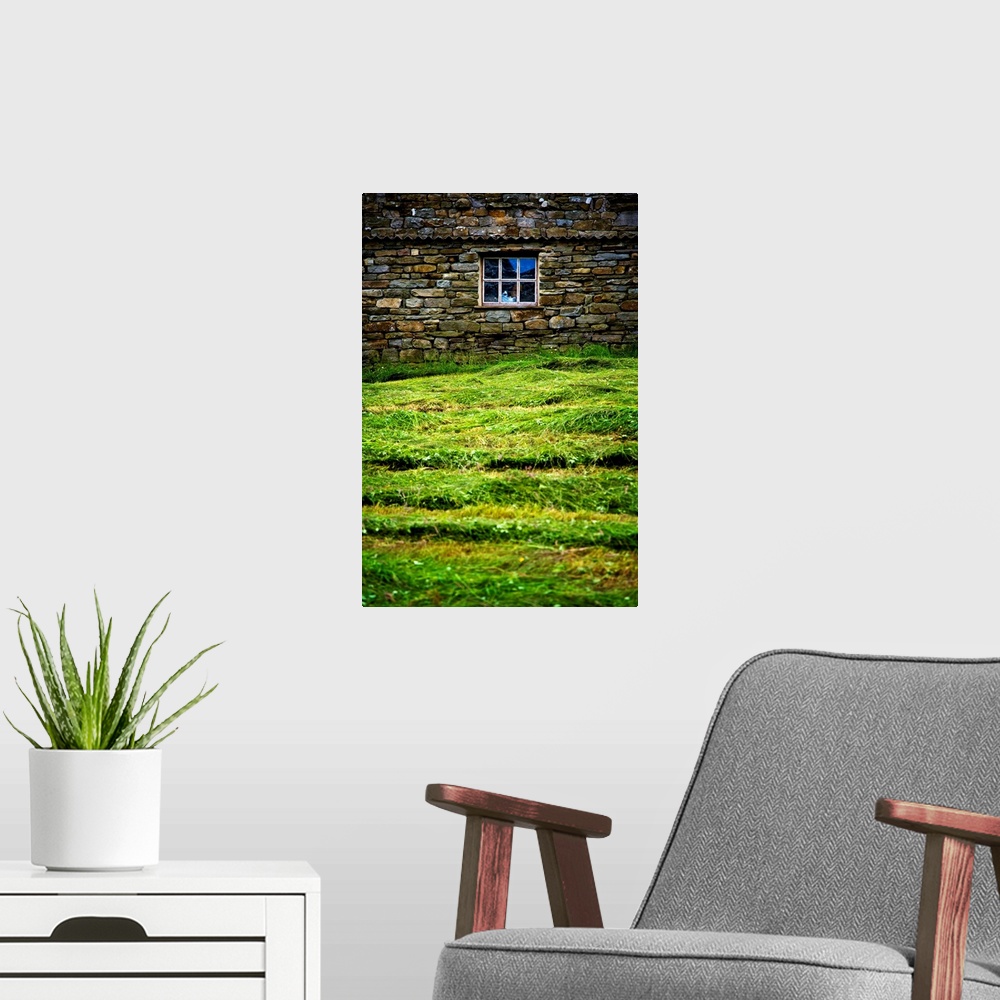 A modern room featuring Photograph taken of a stone wall with a window in it and thick cut grass just in front.