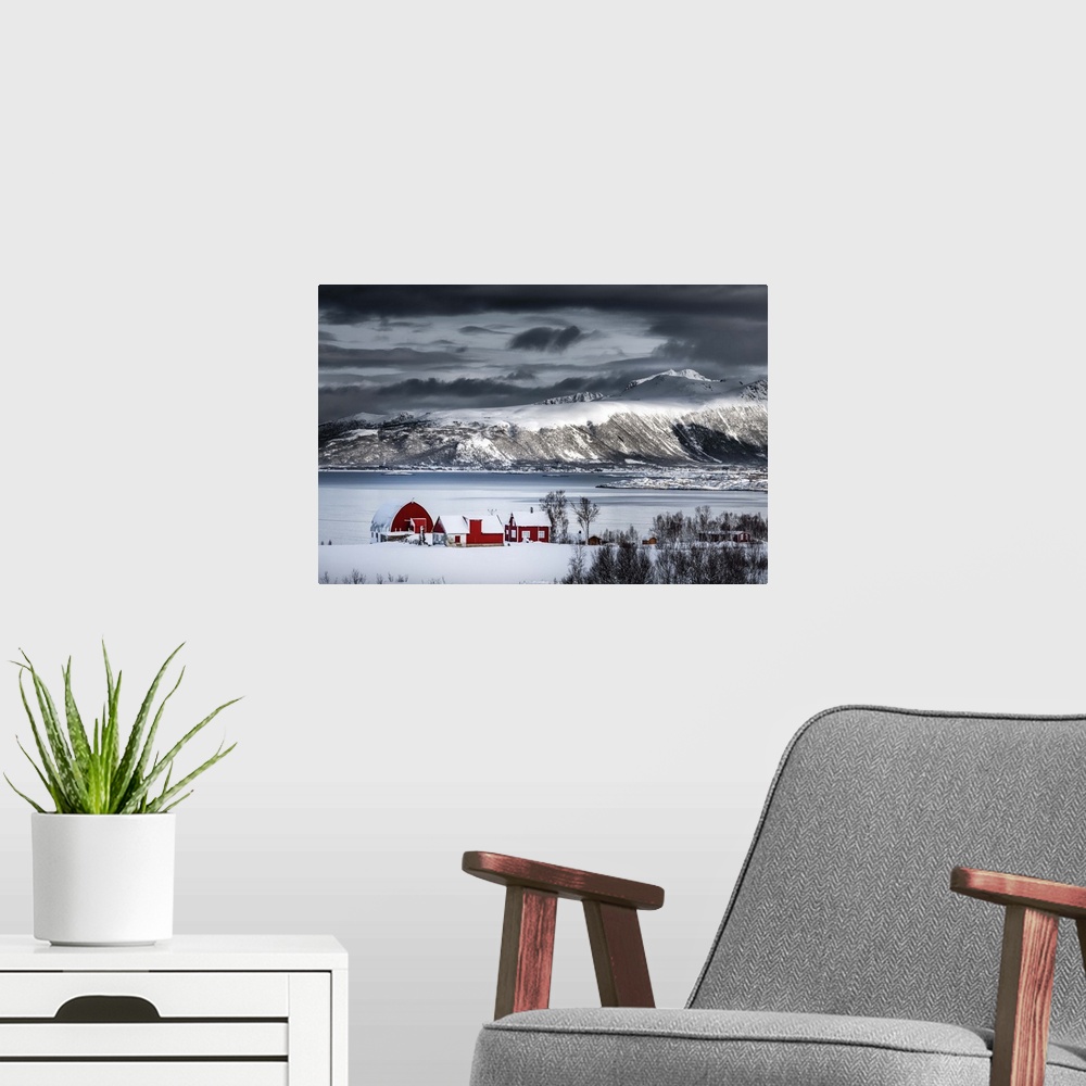 A modern room featuring A photograph of a red building sitting on the shoreline of a snowy landscape with mountains in th...