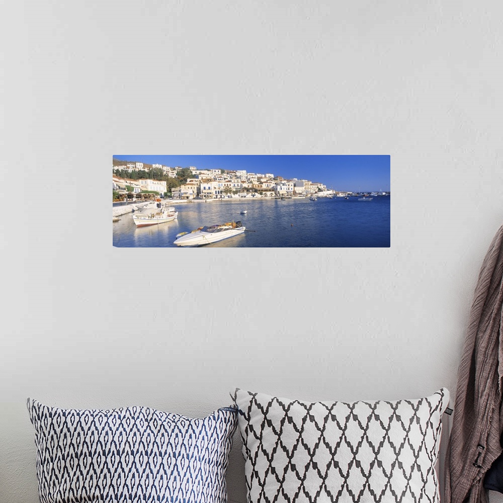 A bohemian room featuring Houses in Greece are photographed from across the water where several boats sit docked.