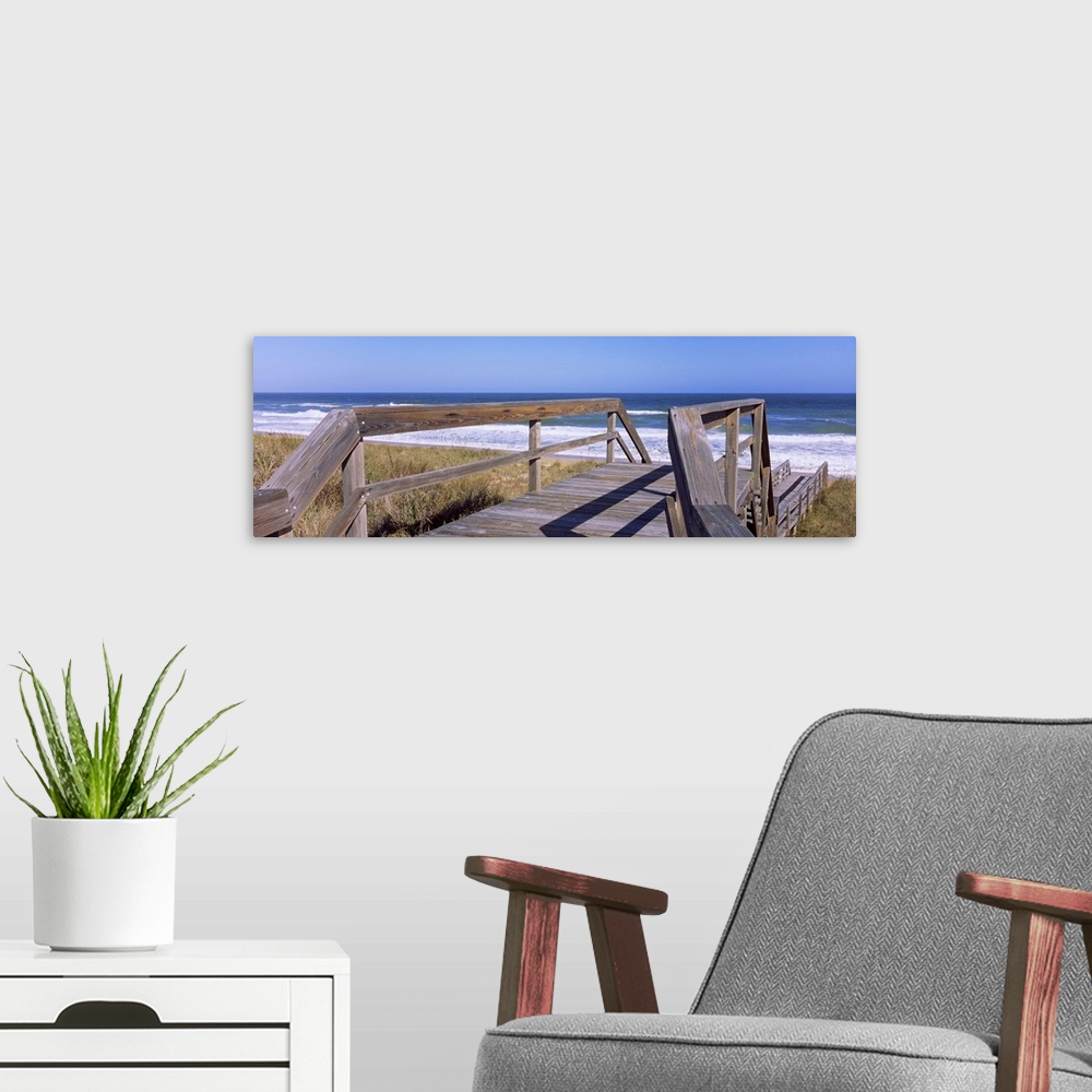 A modern room featuring Panoramic picture taken of a small walking bridge headed down toward the beach with dunes on eith...