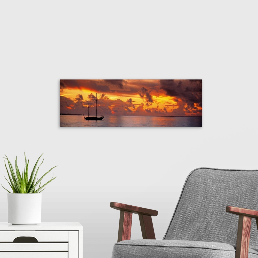 A modern room featuring Single ship with two tall masts on calm waters against a backdrop of dramatic fiery clouds at dusk.