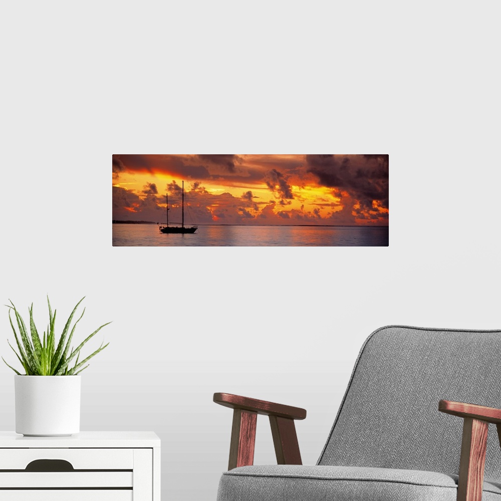 A modern room featuring Single ship with two tall masts on calm waters against a backdrop of dramatic fiery clouds at dusk.