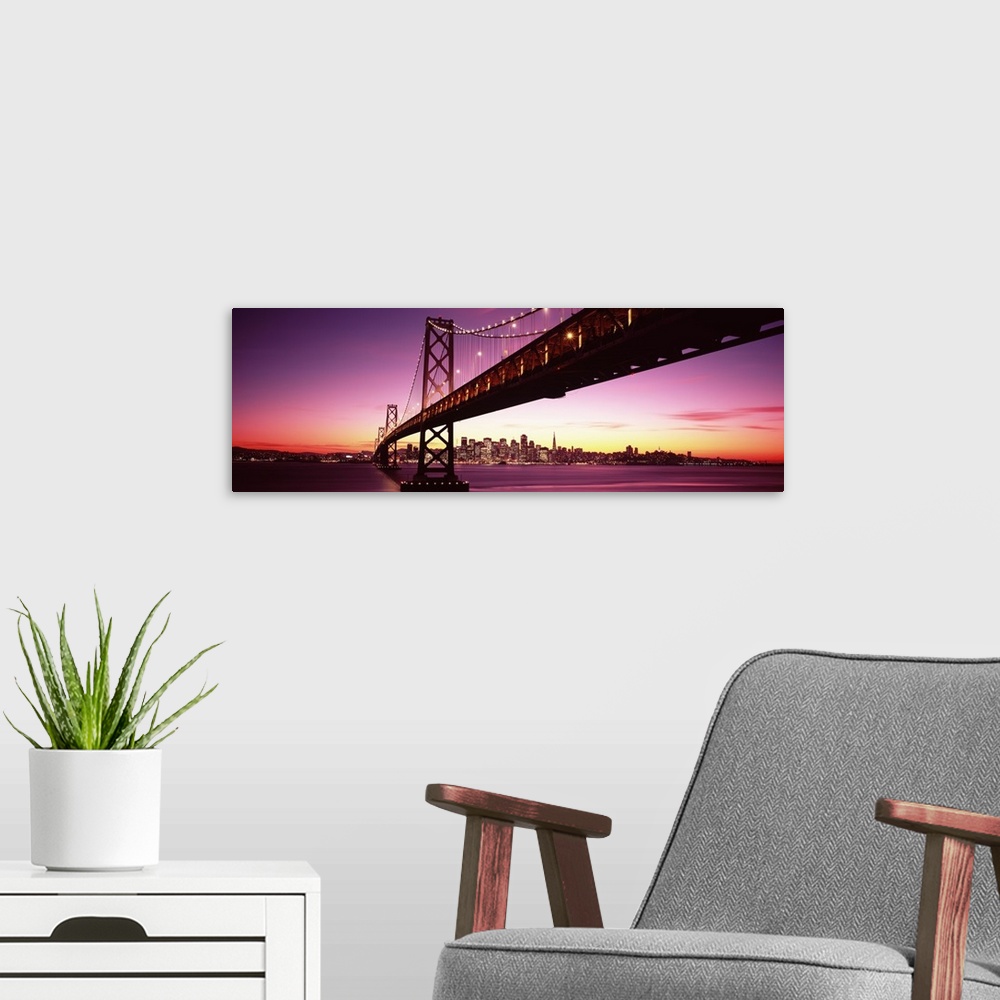 A modern room featuring Large panoramic photo print of a long bridge leading to a lit up city off in the distance at sunset.