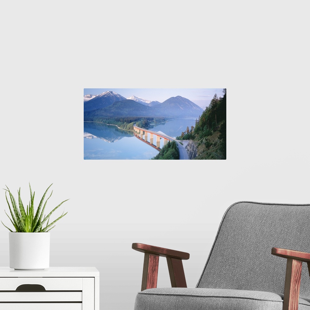 A modern room featuring A winding bridge is photographed as it crosses a large lake into thick forest with a mountain ran...