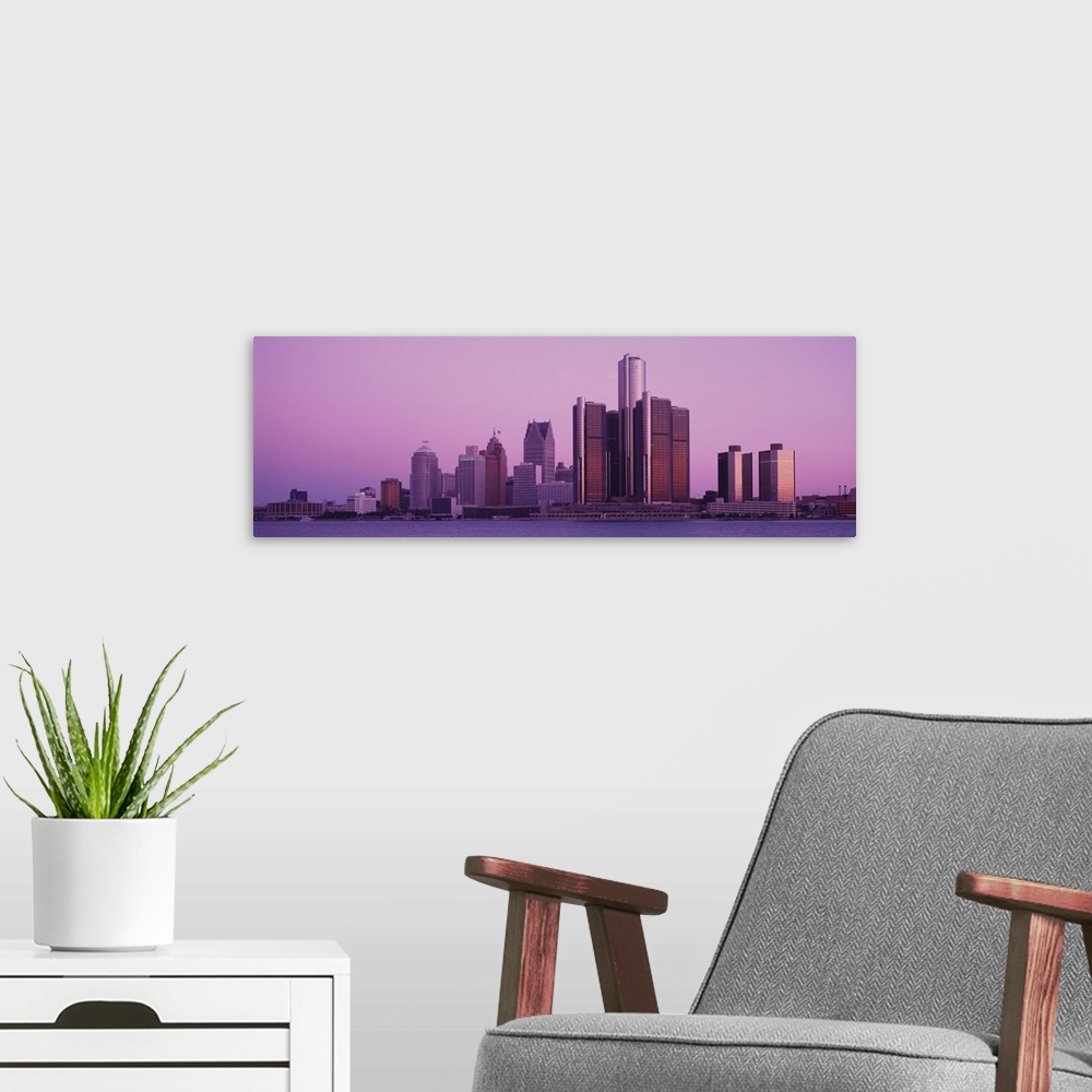 A modern room featuring The Detroit skyline is photographed in panoramic view from across the waterfront during dusk.