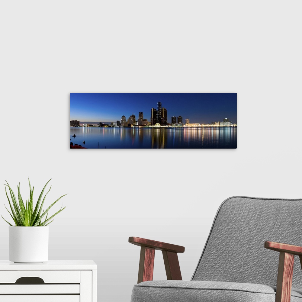 A modern room featuring Narrow canvas print of the Detroit skyline illuminated in the evening with it's lights reflecting...