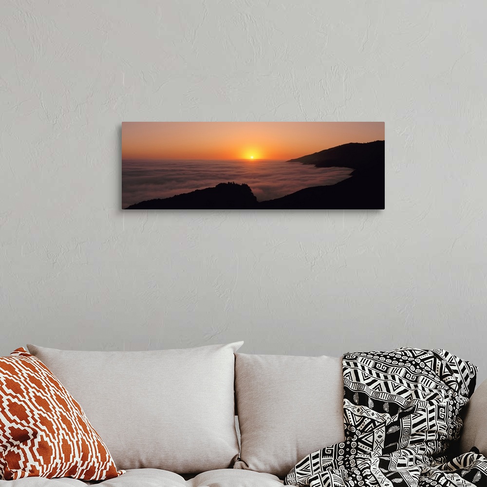 A bohemian room featuring A large panoramic piece of a sunset with a fog mist over the water and mountains lining the right...