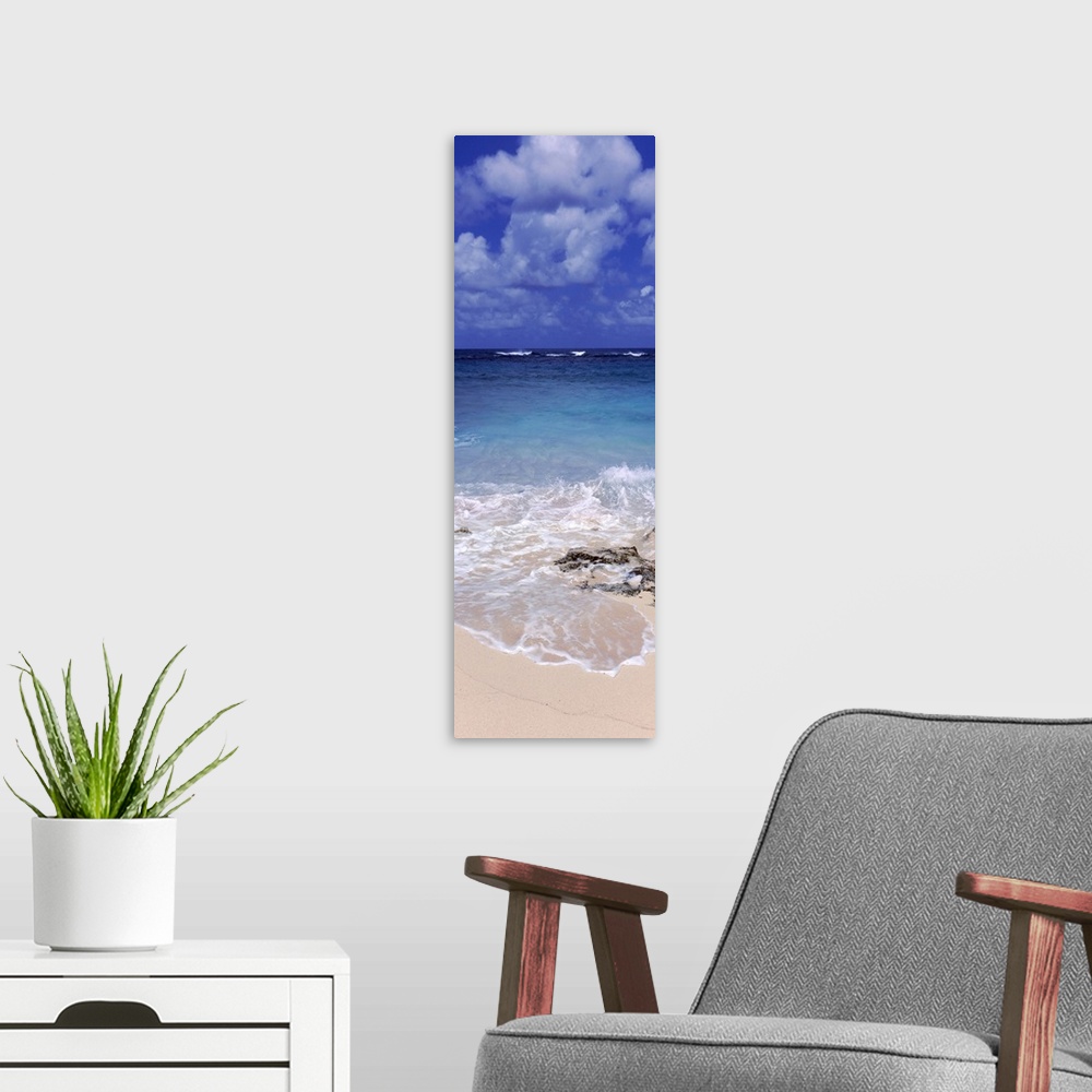 A modern room featuring Vertical, large photograph of clear blue waters hitting the beach beneath a blue sky with billowi...