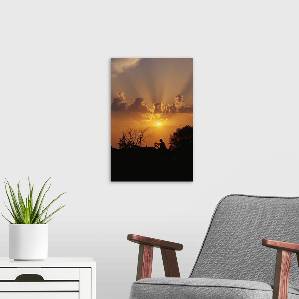 A modern room featuring This large vertical piece shows a farmer driving a tractor that is silhouetted by the sunset behi...